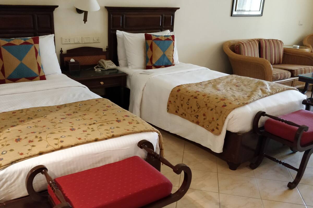 A hotel room with two beds, a wooden headboard, a bedside table with a lamp, a chair, and a small table with a telephone.