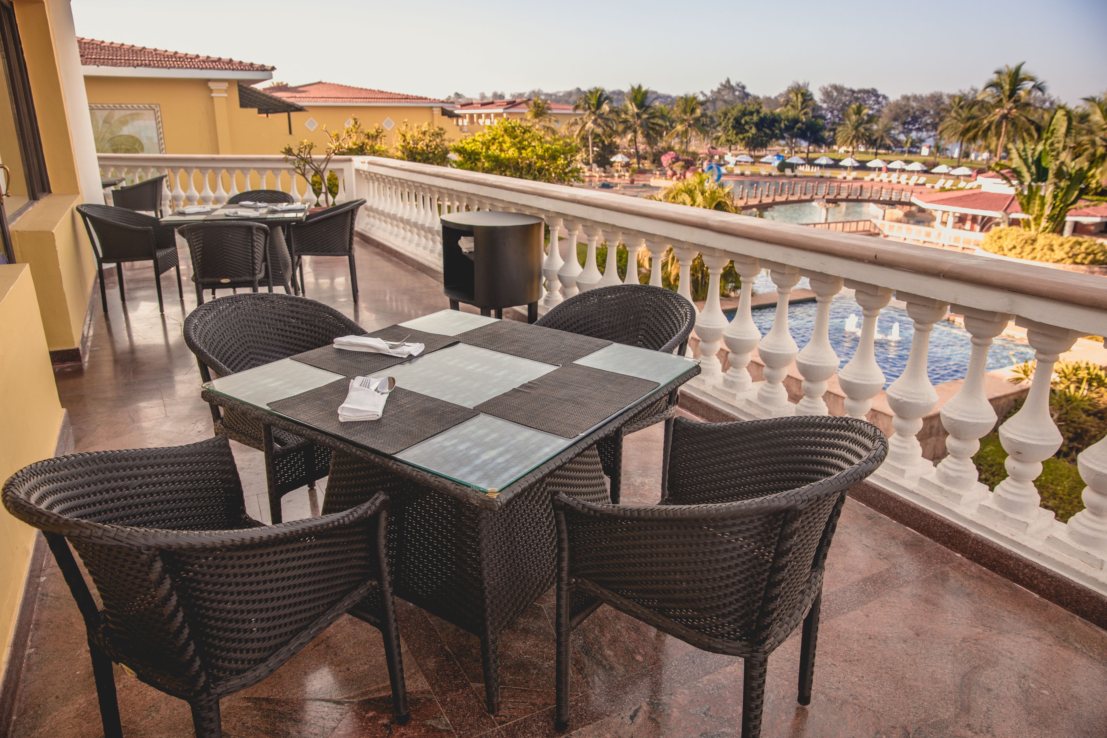 A balcony with wicker furniture and a glass table, overlooking a pool and palm trees.