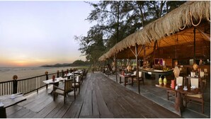 A wooden deck with tables and chairs, overlooking a beach at sunset.