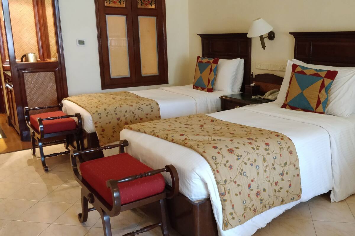 A hotel room with two beds, each with a floral bedspread and matching pillows. There is a wooden chair with a red cushion and a small table with a lamp.