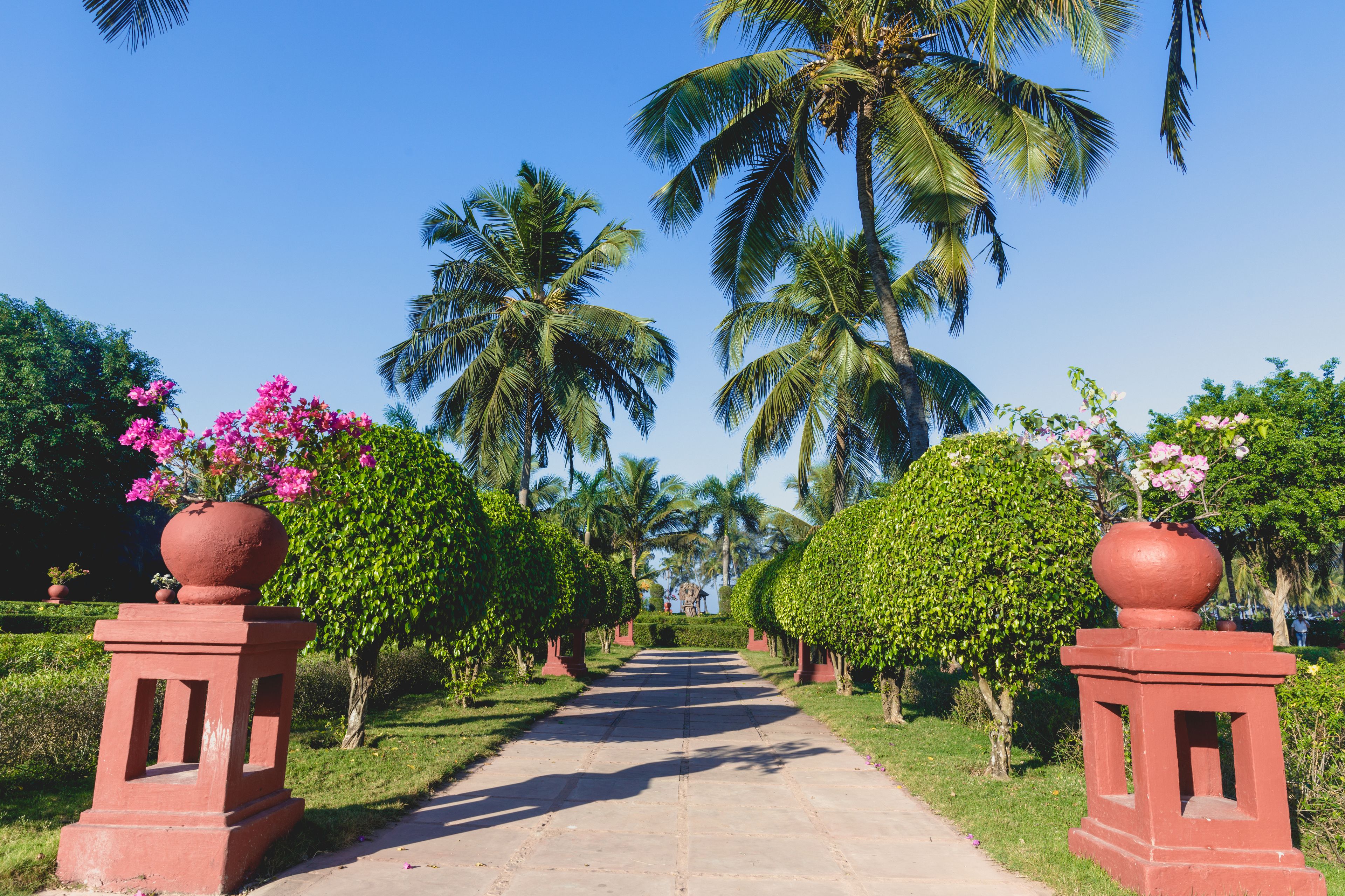 A pathway flanked by red pillars with spherical tops, leading towards palm trees and a clear blue sky.