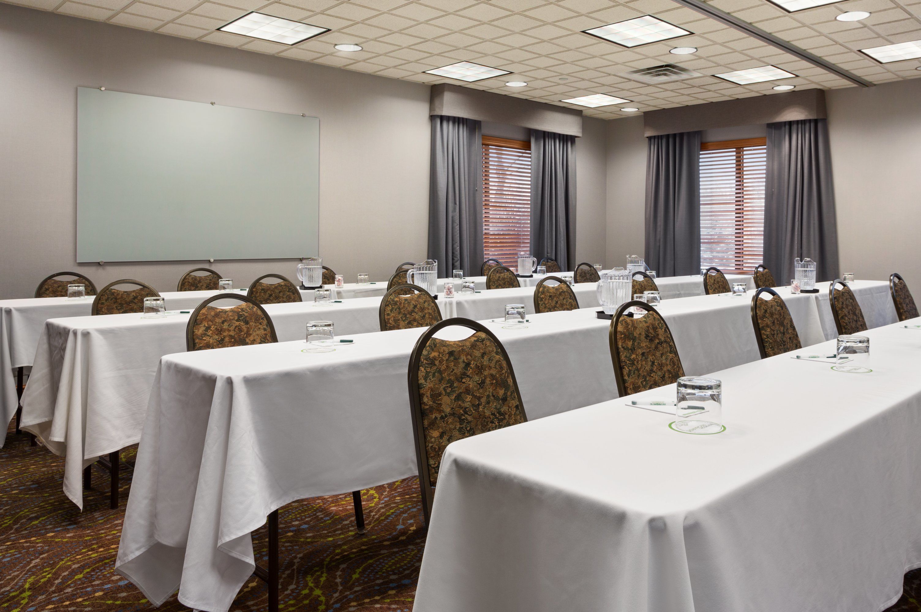 A conference room with multiple tables, chairs, a projector screen, and windows with curtains.