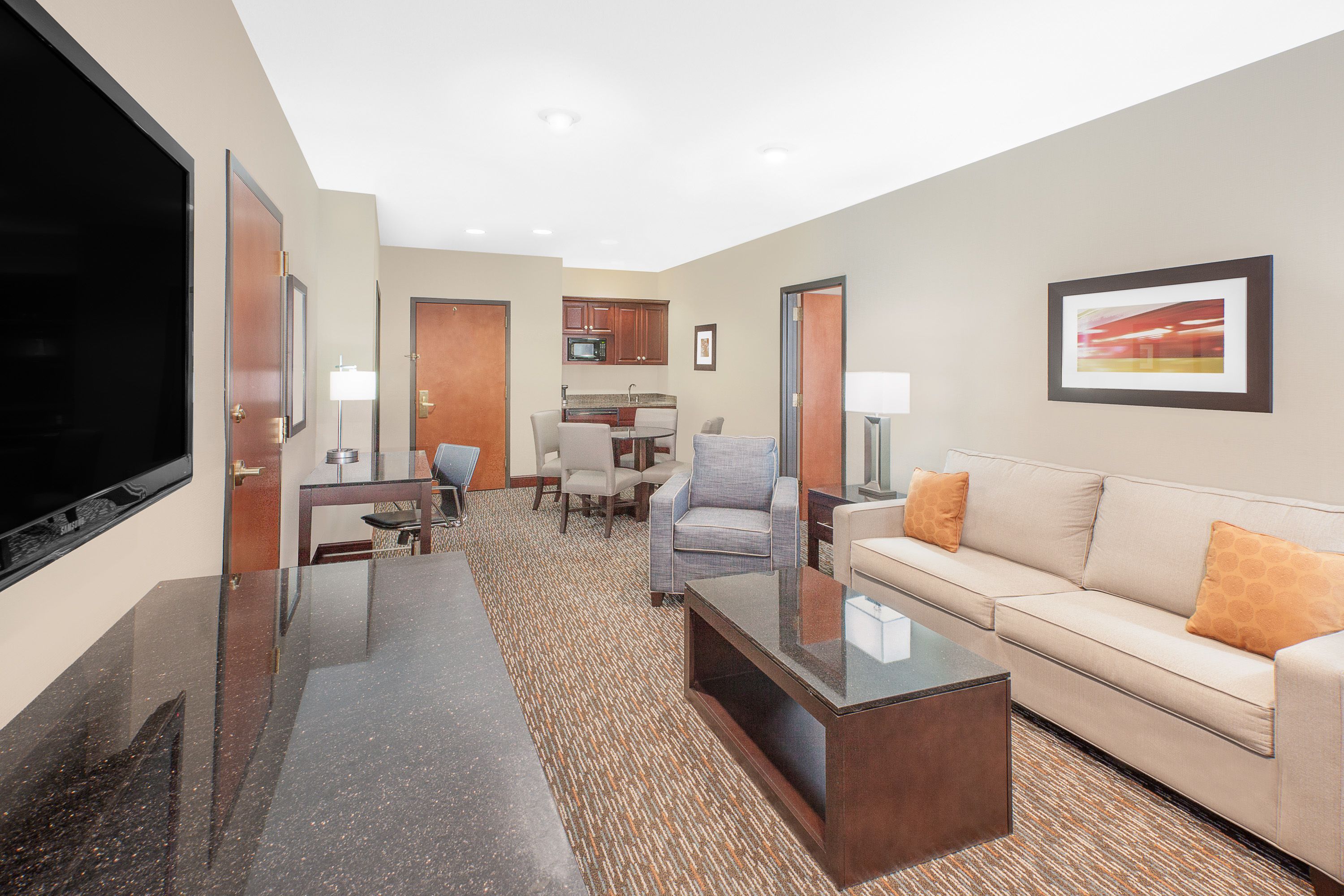 Habitación de hotel con televisión de pantalla plana, sofá, mesa de centro, zona de comedor con sillas y cocina equipada con armarios y microondas.