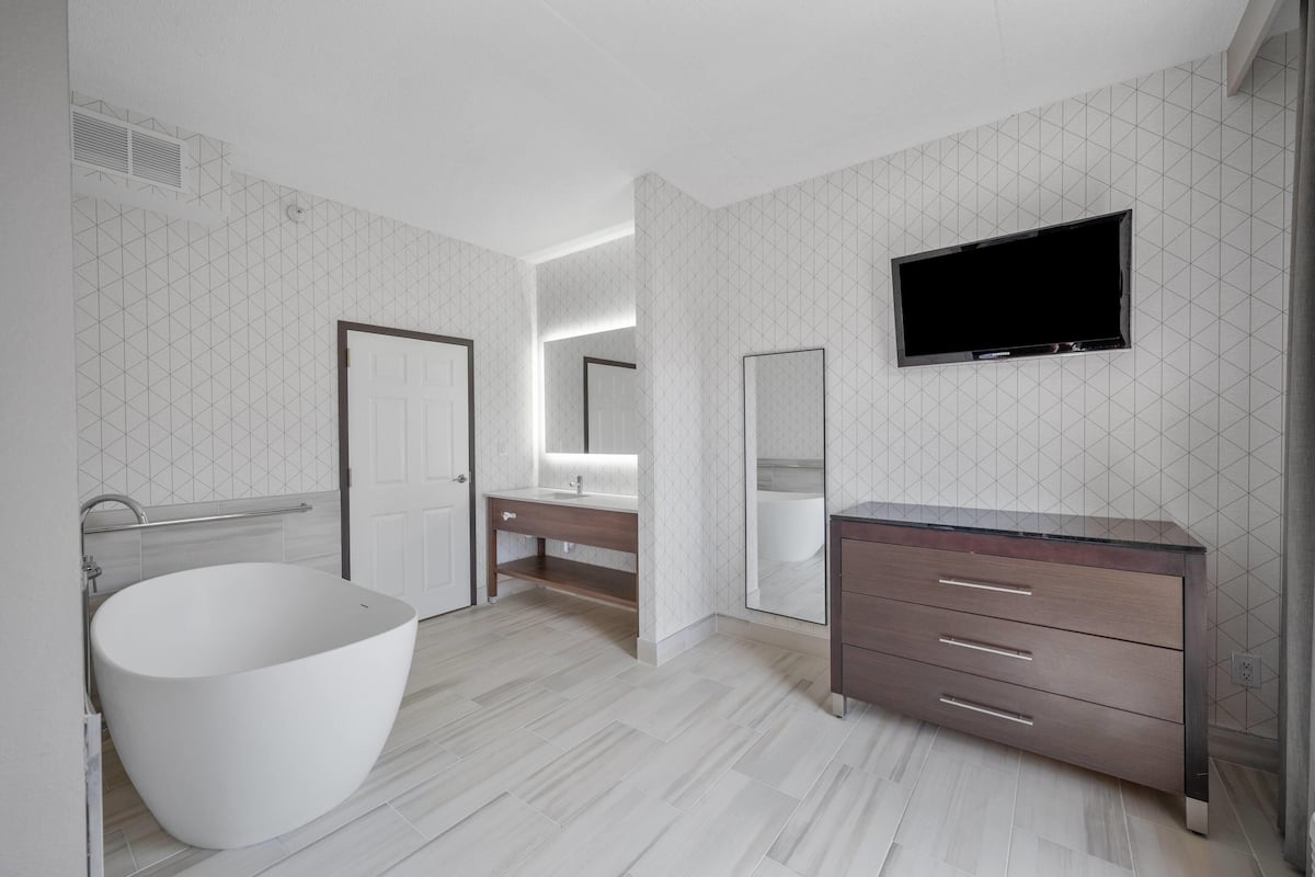 A modern bathroom with a freestanding bathtub, a vanity with a mirror, and a flat-screen TV mounted on the wall.