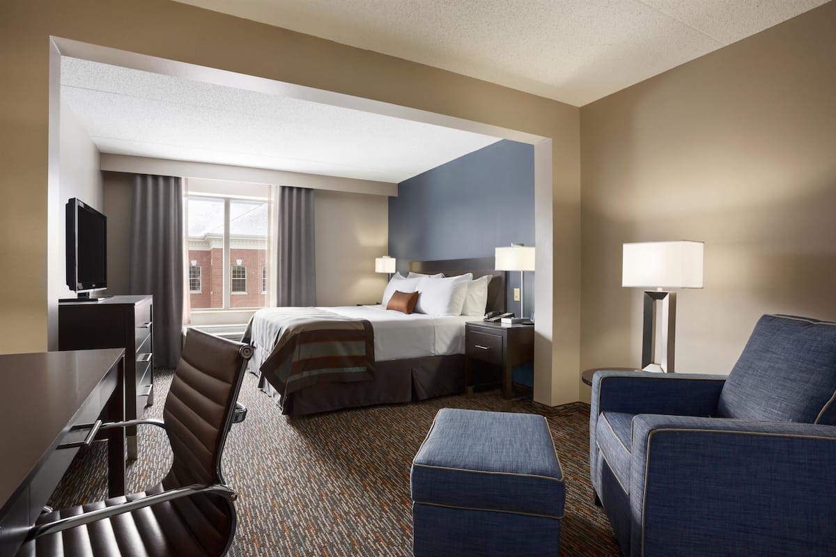 A hotel room with a bed, desk, chair, ottoman, lamps, and a view of a building through the window.