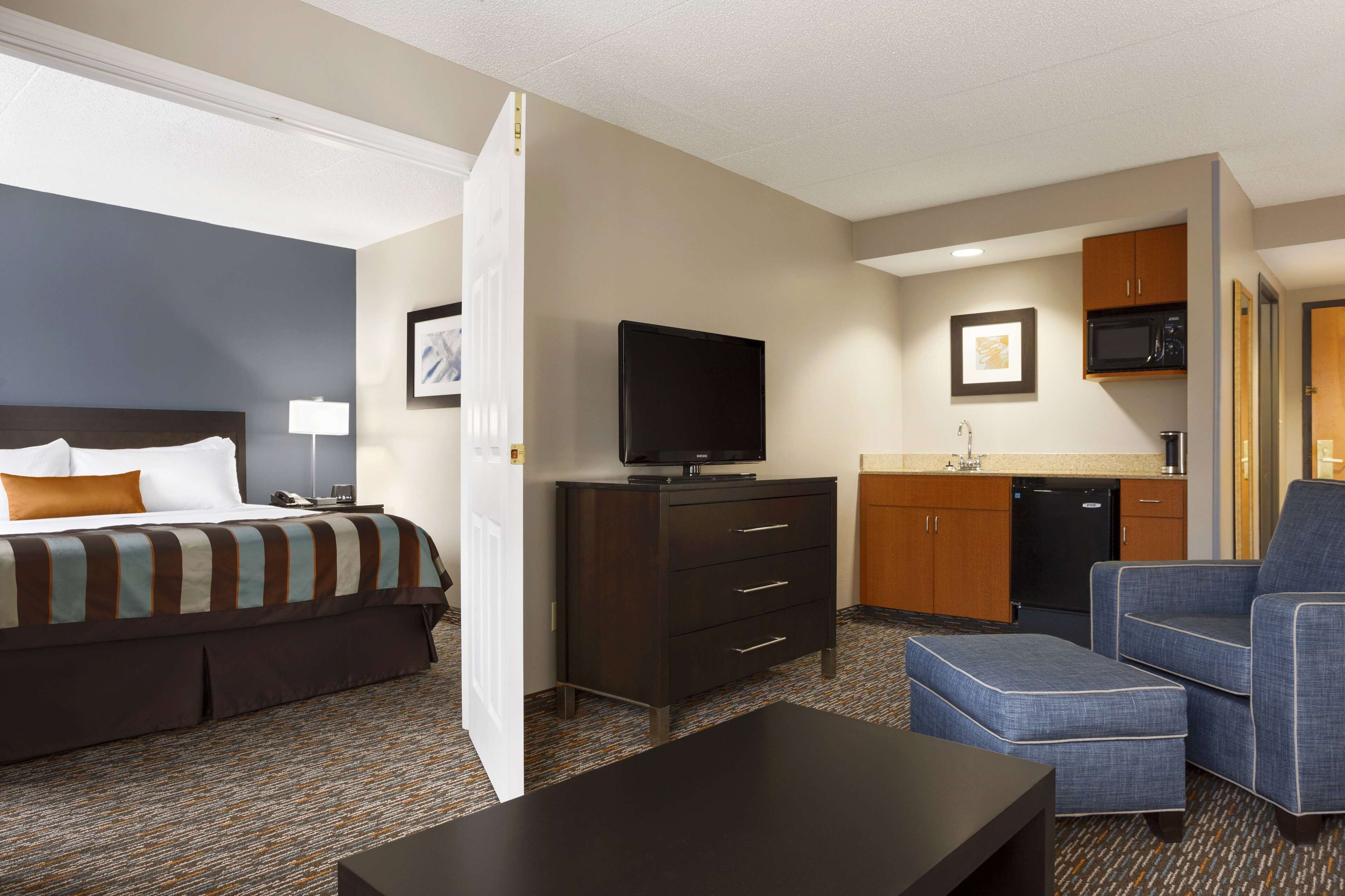 A hotel room with a bed, a TV, a desk, a chair, a kitchenette, and a bathroom.
