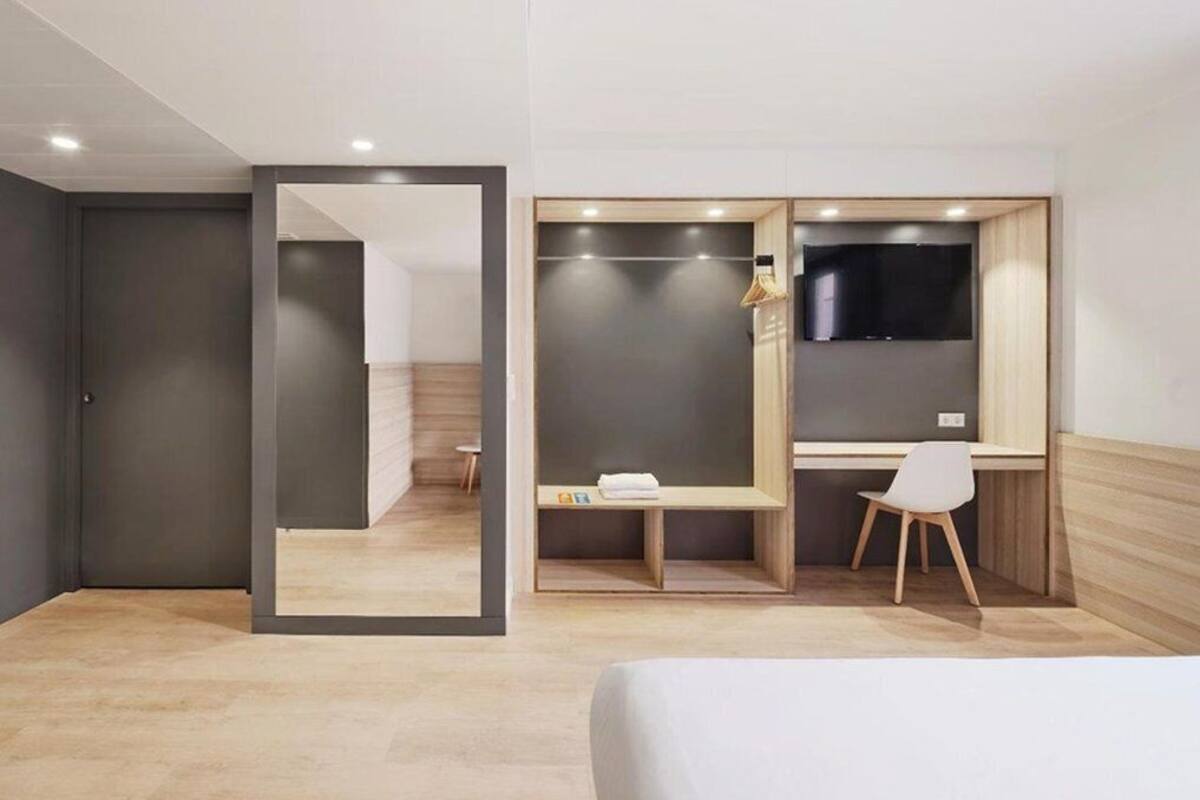 A modern hotel room with a bed, a desk, a chair, a TV, and a wardrobe.