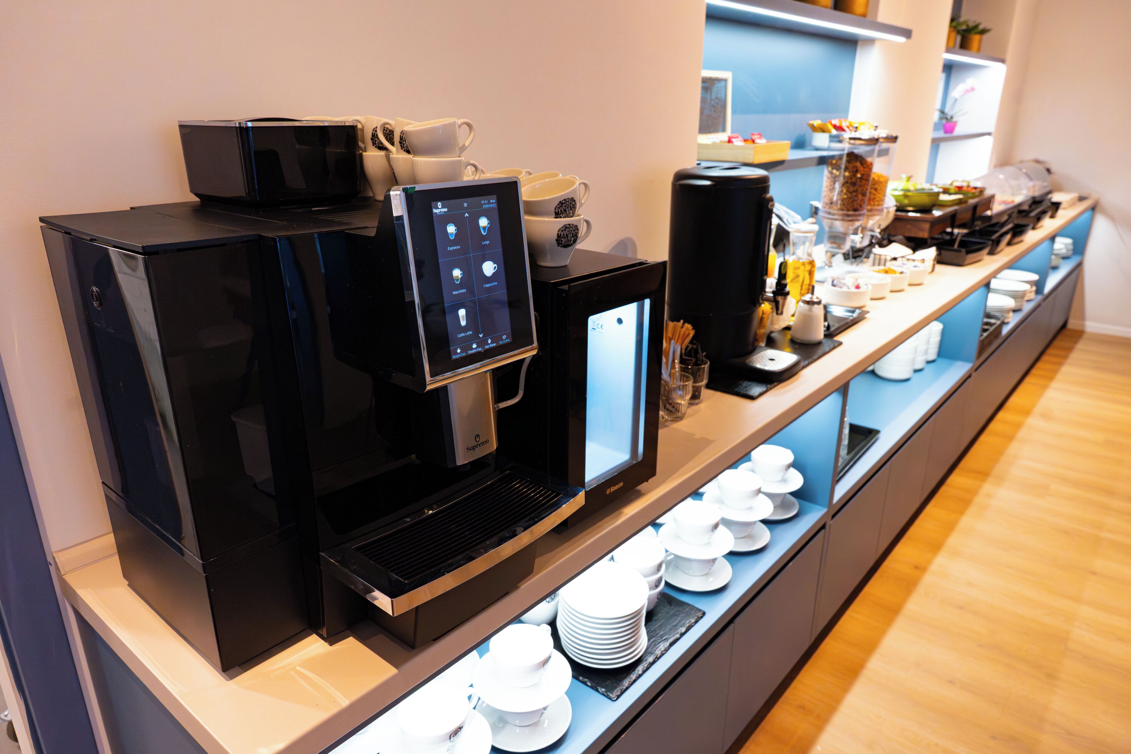 A coffee station with a digital menu, coffee machines, and a variety of coffee cups and bowls.