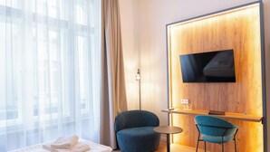 A hotel room with a bed, a desk with a chair, a TV mounted on a wooden wall, and a large window with sheer curtains. - Pilvax Hotel Budapest (Budapest)