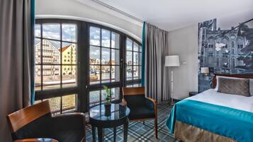 Ein Hotelzimmer mit großem Fenster, einem Bett, einem Sessel, einem Tisch und einer Lampe.