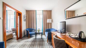 Ein Hotelzimmer mit einem Bett, zwei blauen Sessel, einem Holztisch mit Fernseher, einem Spiegel und einem gemusterten Teppich.