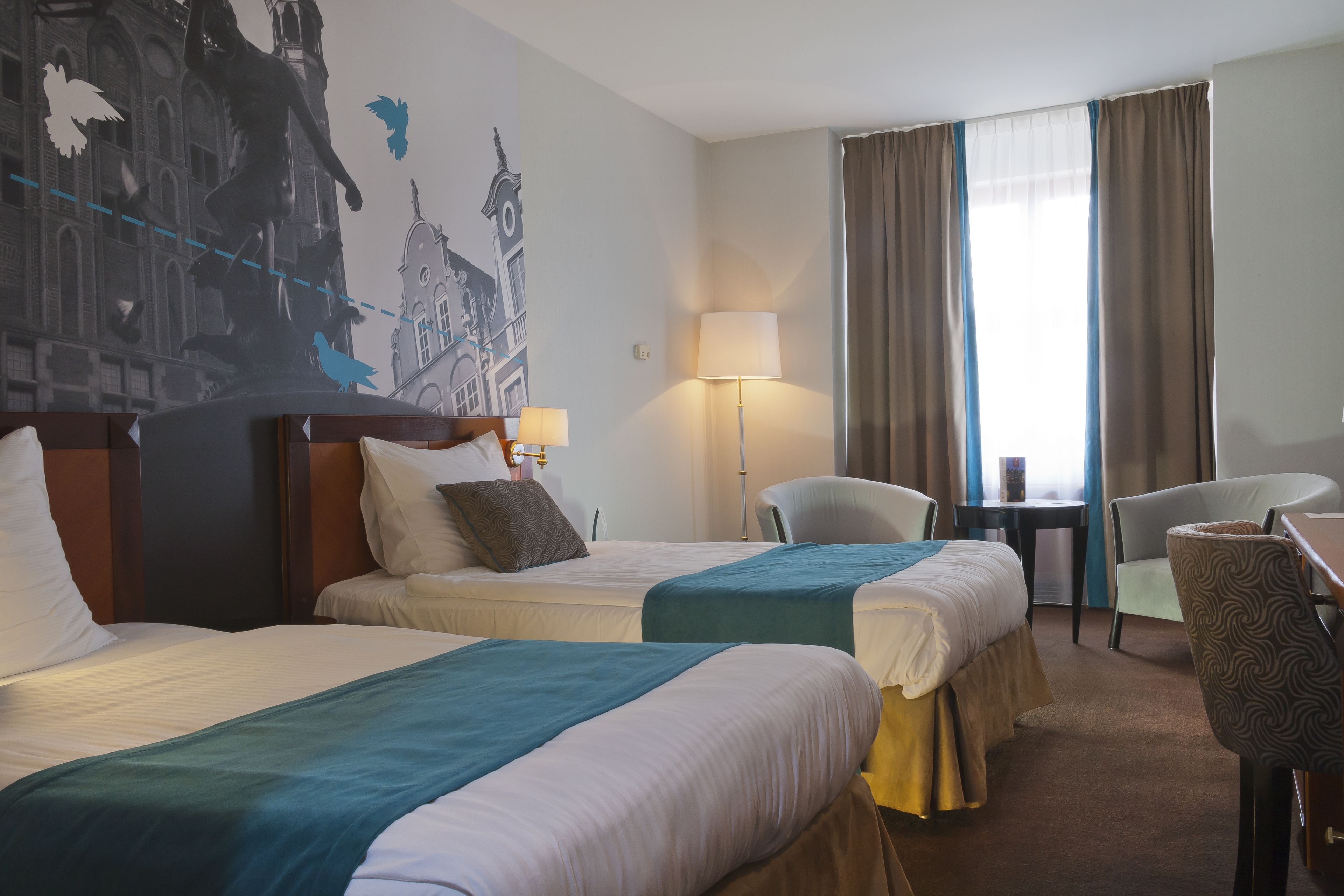 A hotel room with two beds, a desk, a chair, a lamp, and a large window with curtains.