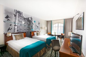 A hotel room with two beds, a desk with a TV, a chair, a lamp, and a large wall mural.