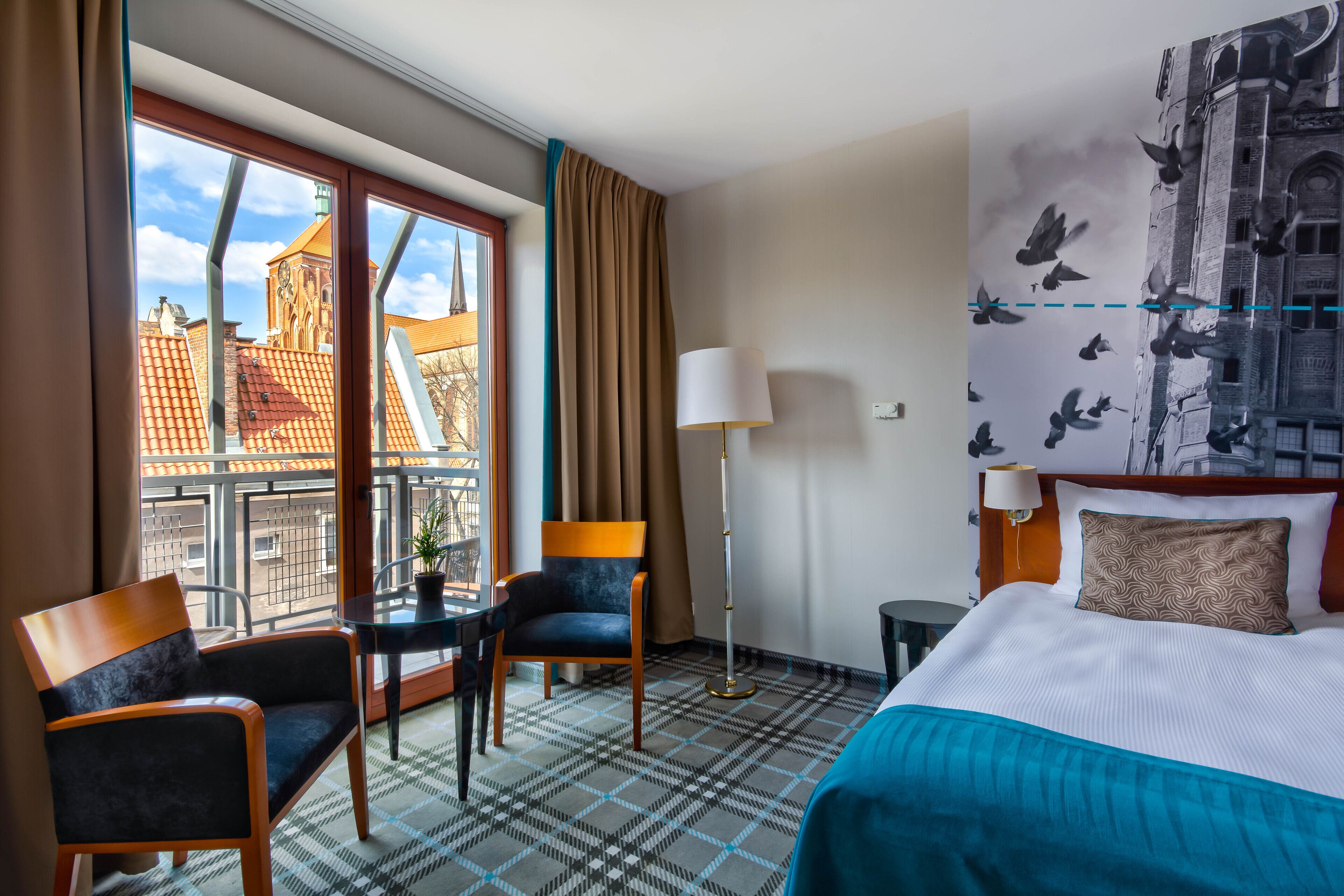 Habitación de hotel con una cama, dos sillas, una mesita, una lámpara y vistas a un edificio por la ventana.
