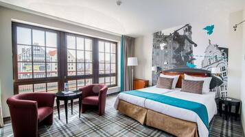 Ein Hotelzimmer mit einem großen Bett, zwei Sesseln, einem kleinen Tisch und Blick auf eine Stadtkulisse durch große Fenster.