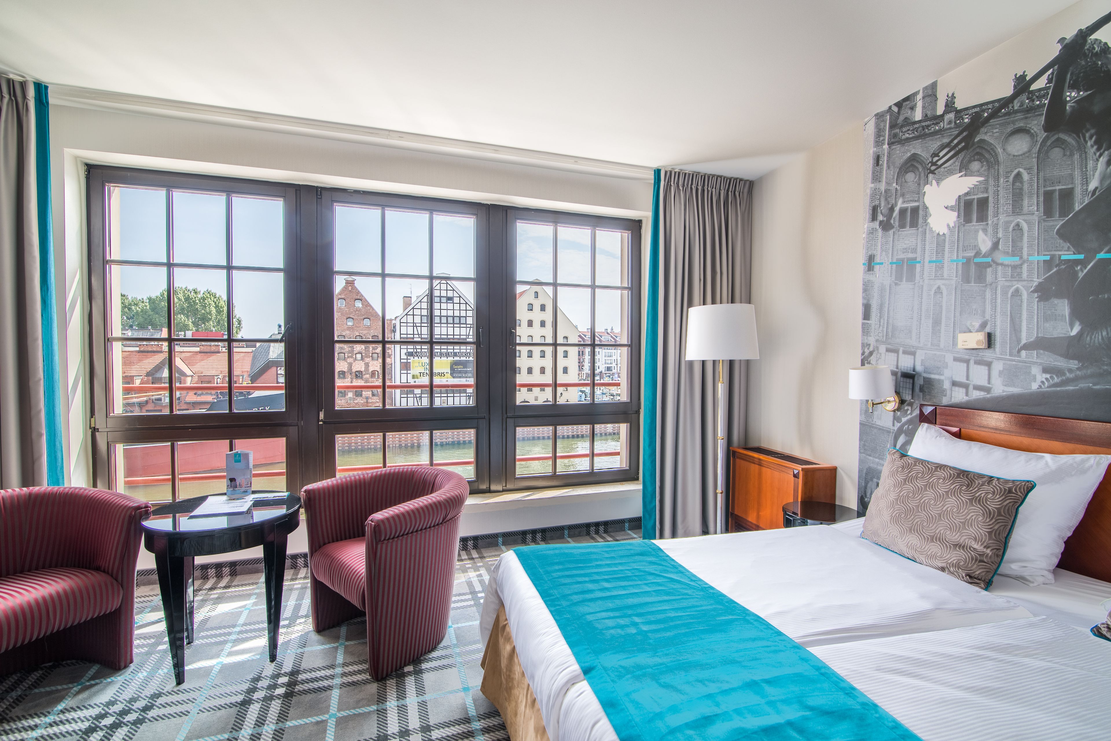 A hotel room with a large window, a bed with a blue blanket, two striped armchairs, a small table, and a lamp.