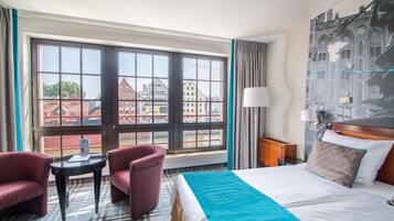 A hotel room with a large window, a bed with a blue blanket, two striped armchairs, a small table, and a lamp.