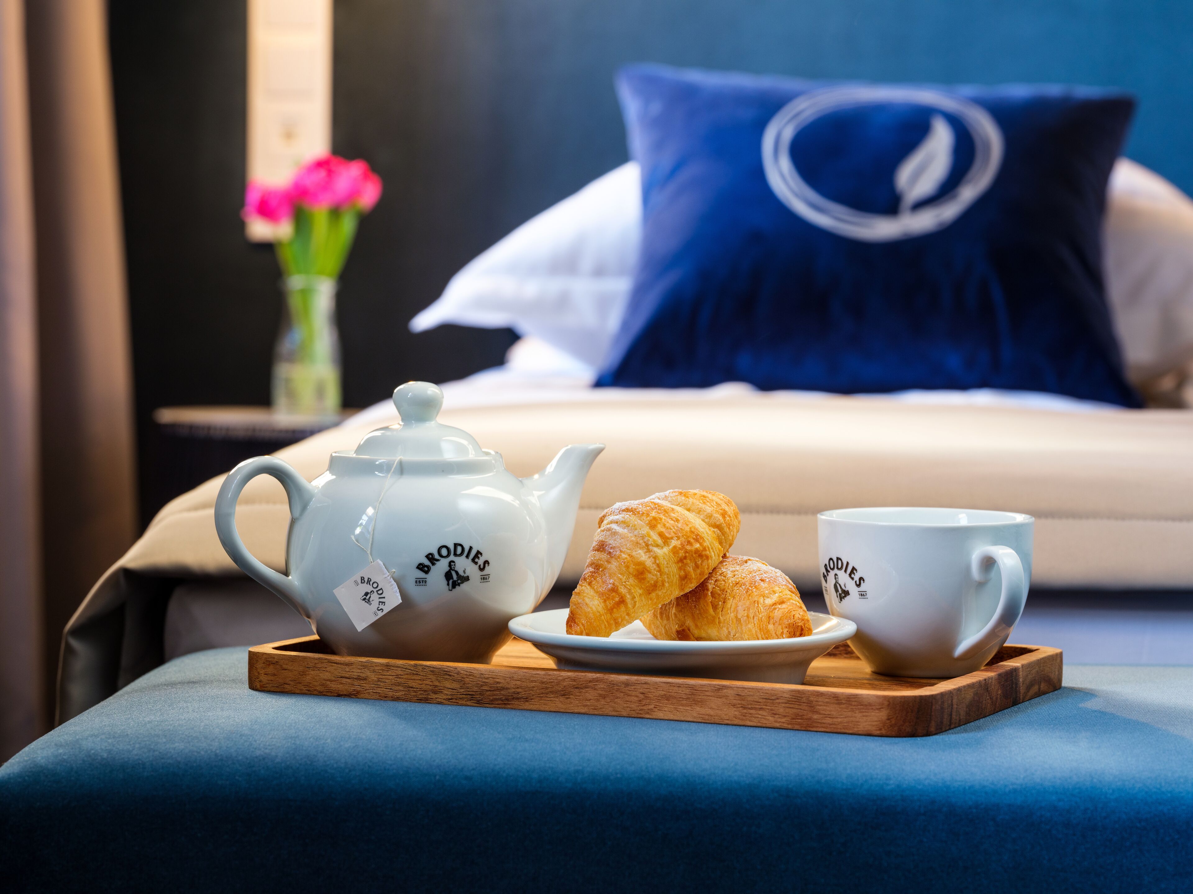 A neatly arranged breakfast setup with a teapot, two cups, and croissants on a tray, placed on a blue upholstered bench in a hotel room.