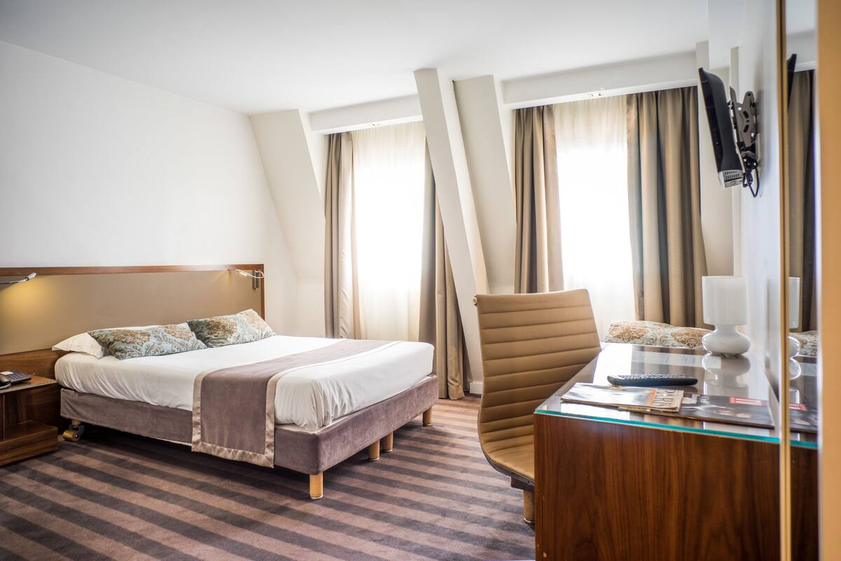 A hotel room with a large bed, a desk with a lamp, a chair, and a TV mounted on the wall.