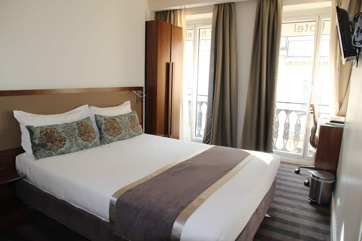 A hotel room with a large bed, a wooden headboard, a small desk, a trash can, and a balcony with curtains.