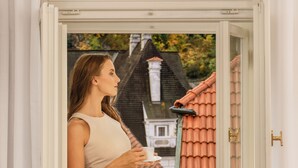 A person standing by a window with a view of a house with a red roof.