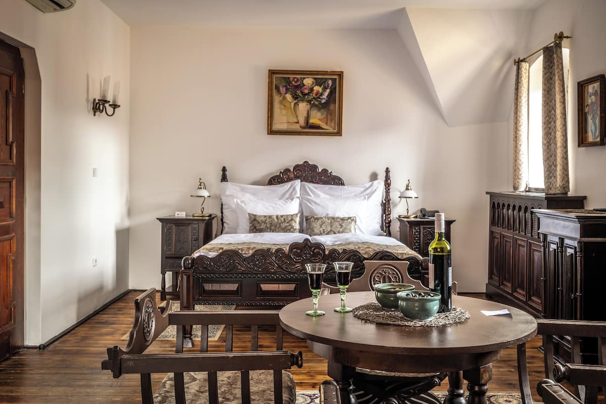 Un dormitorio con una cama grande, una mesa redonda de madera con dos vasos y una botella, un cuadro en la pared y un armario de madera.