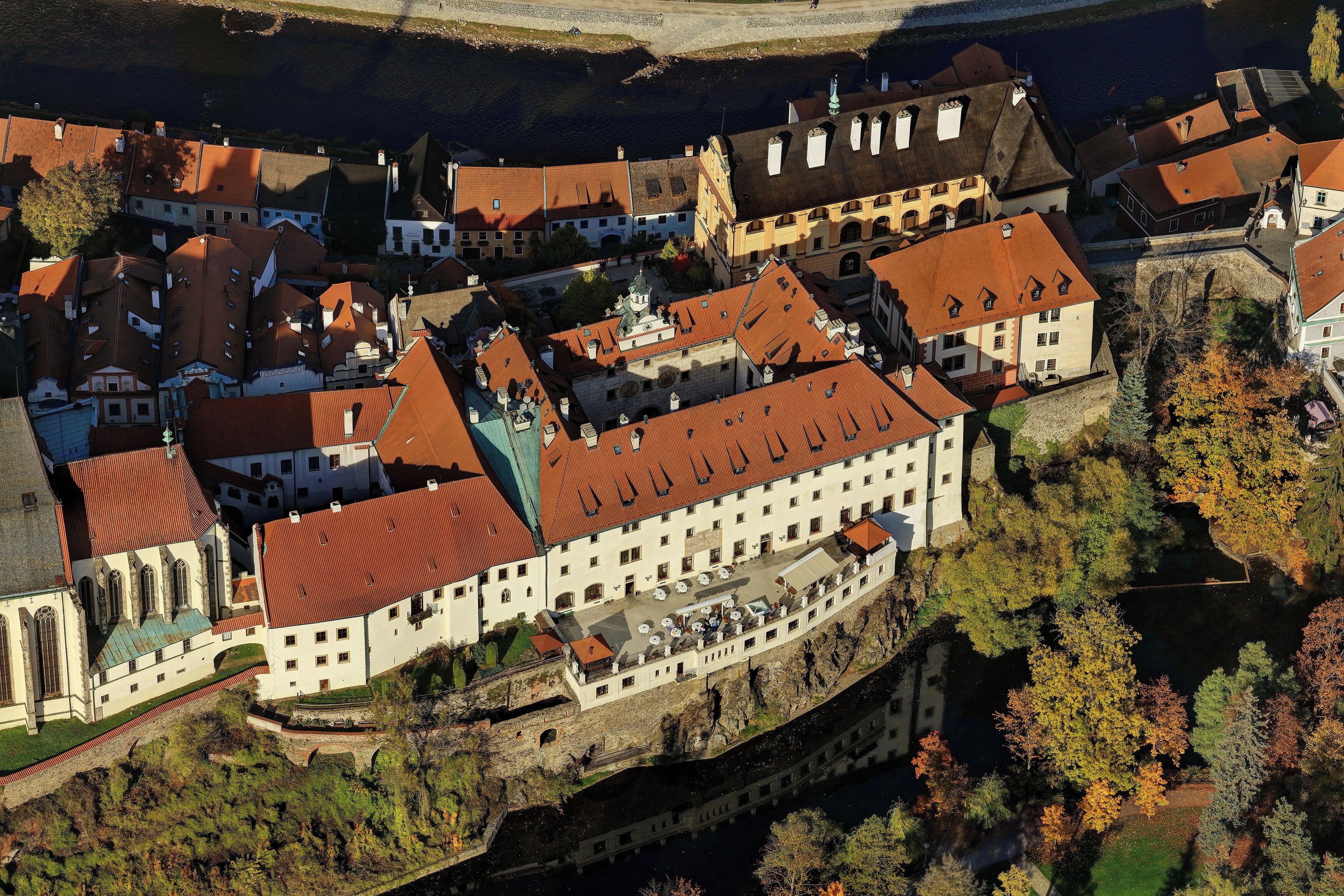 An aerial view of a historic town with a prominent castle-like building surrounded by smaller structures, a river, and autumn foliage.