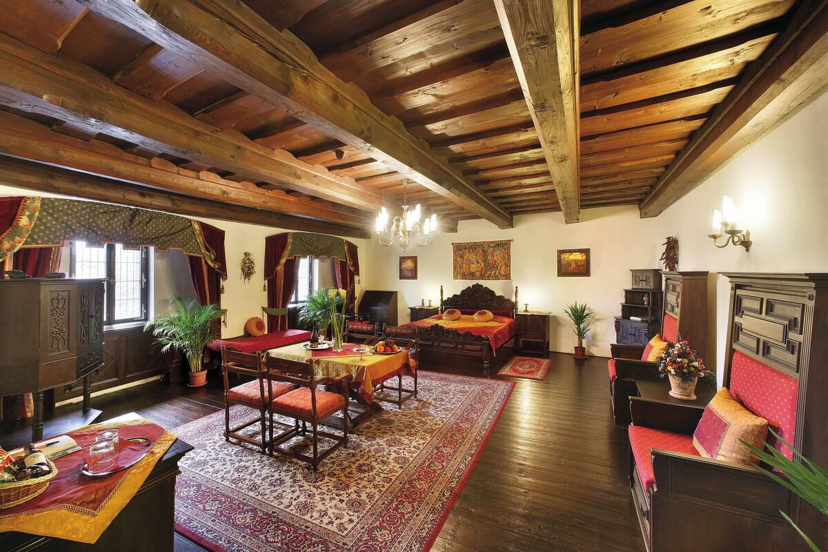 Una habitación tradicional con vigas de madera, una chimenea, una cama, una mesa de comedor con sillas y un sofá rojo.