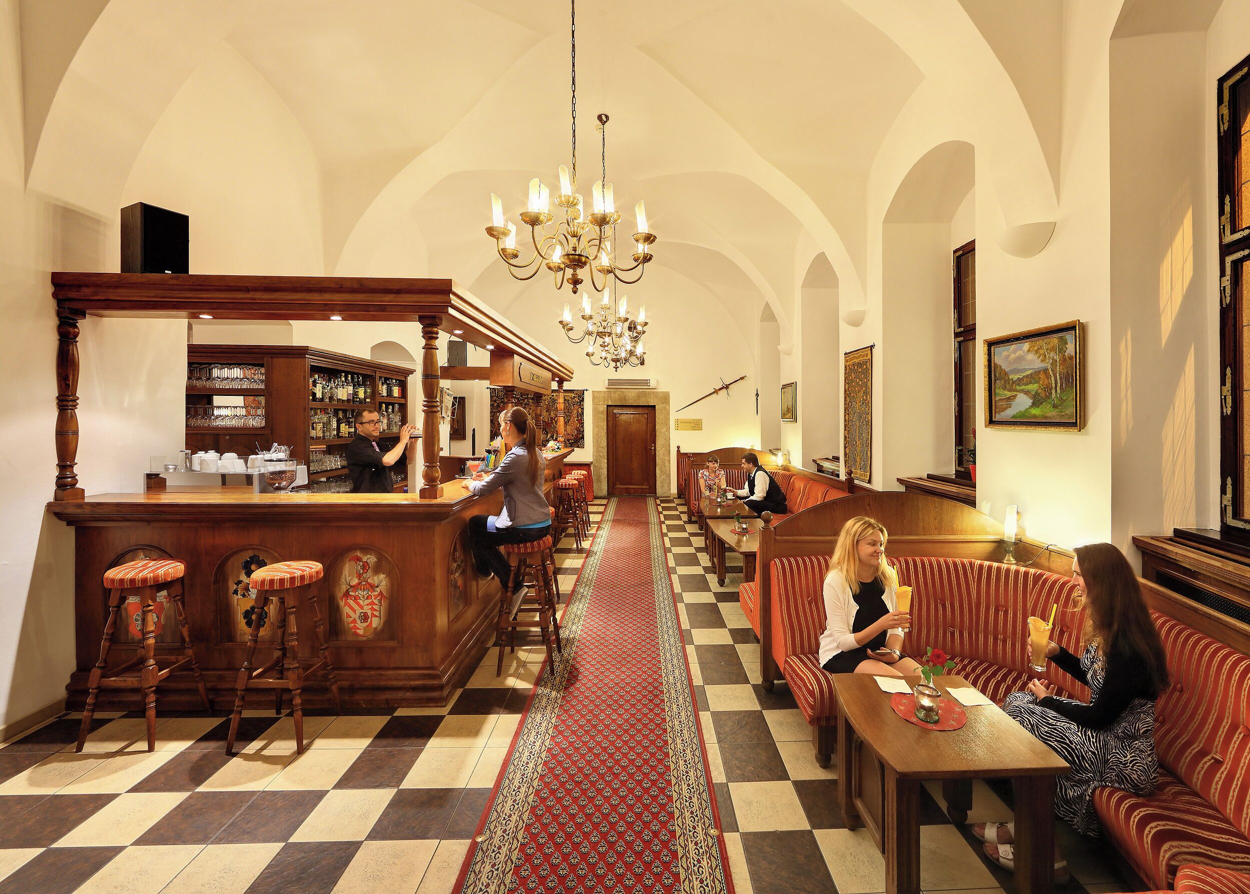 A bar area with a checkered floor, wooden bar counter, and stools. There are seating areas with patterned cushions and a chandelier.