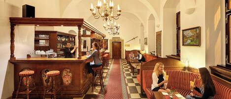 A bar area with a checkered floor, wooden bar counter, and stools. There are seating areas with patterned cushions and a chandelier.