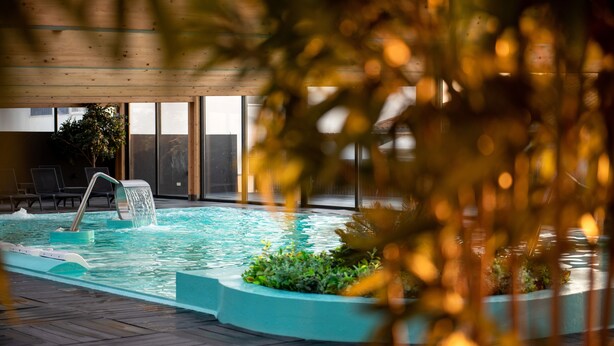 An indoor swimming pool with a water feature, surrounded by a wooden deck and glass walls.