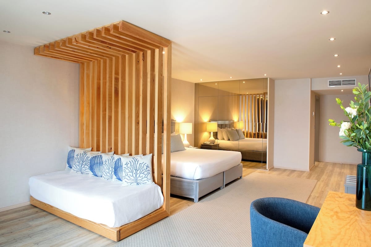 Ein modernes Hotelzimmer mit einem Holz-Kopfteil, einem Bett mit weißer Bettdecke, einem blau gepolsterten Sessel und einem Holztisch.