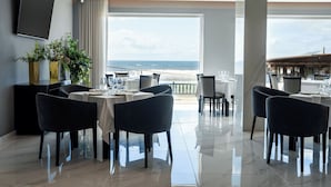 A modern dining area with a view of the ocean, featuring a large window, black chairs, and a flat-screen TV mounted on the wall.