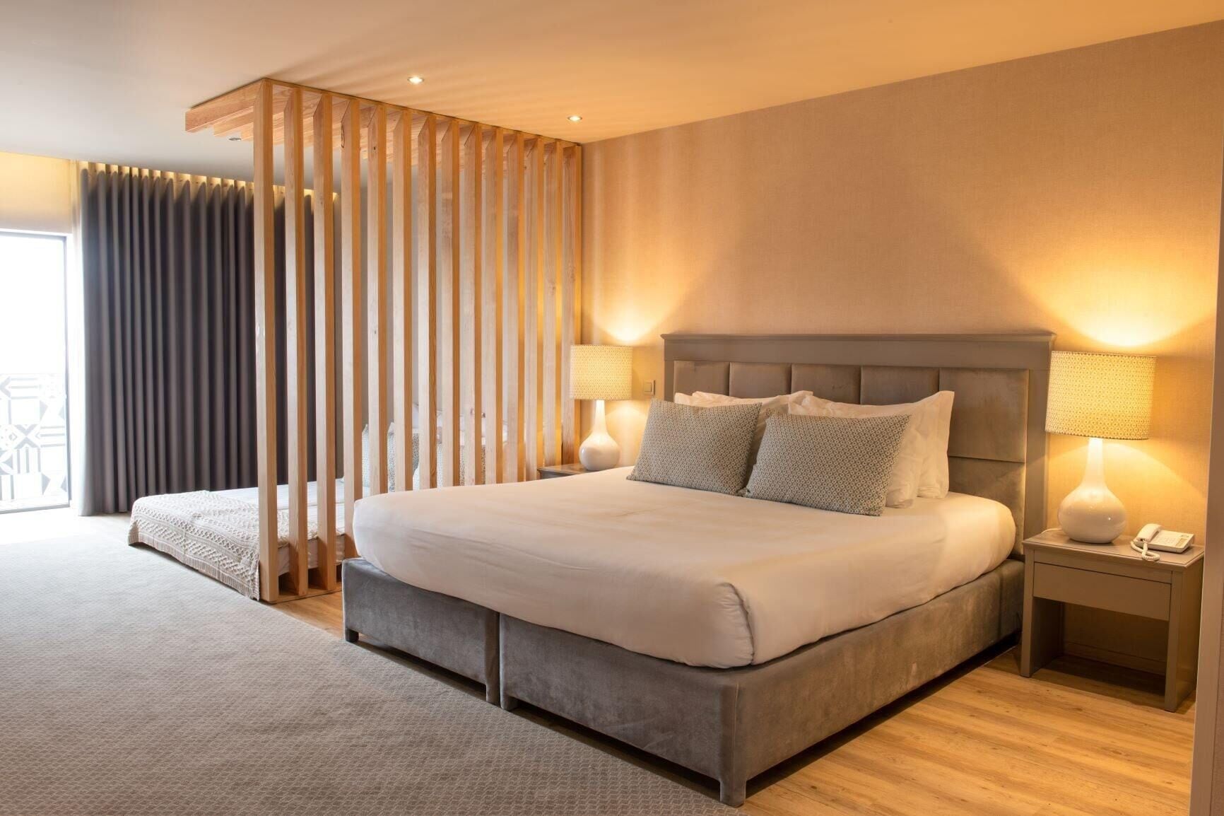 A modern hotel room with a large bed, two bedside tables, and a view through a window with curtains.