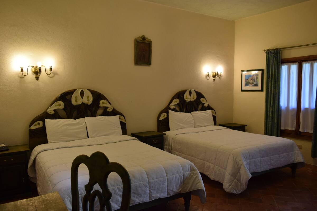 A room with two beds, each with a wooden headboard and white bedding. There are wall-mounted light fixtures on either side of the beds.