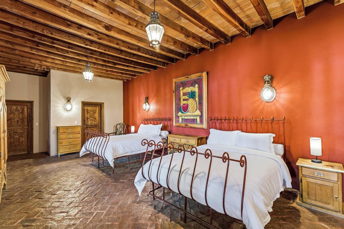 A room with two beds, wooden beams on the ceiling, a red wall, and a fruit-themed painting.