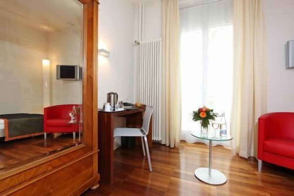 Une pièce avec un plancher en bois, une chaise rouge, une table en verre ornée d’un arrangement floral, une chaise blanche et un meuble en bois.