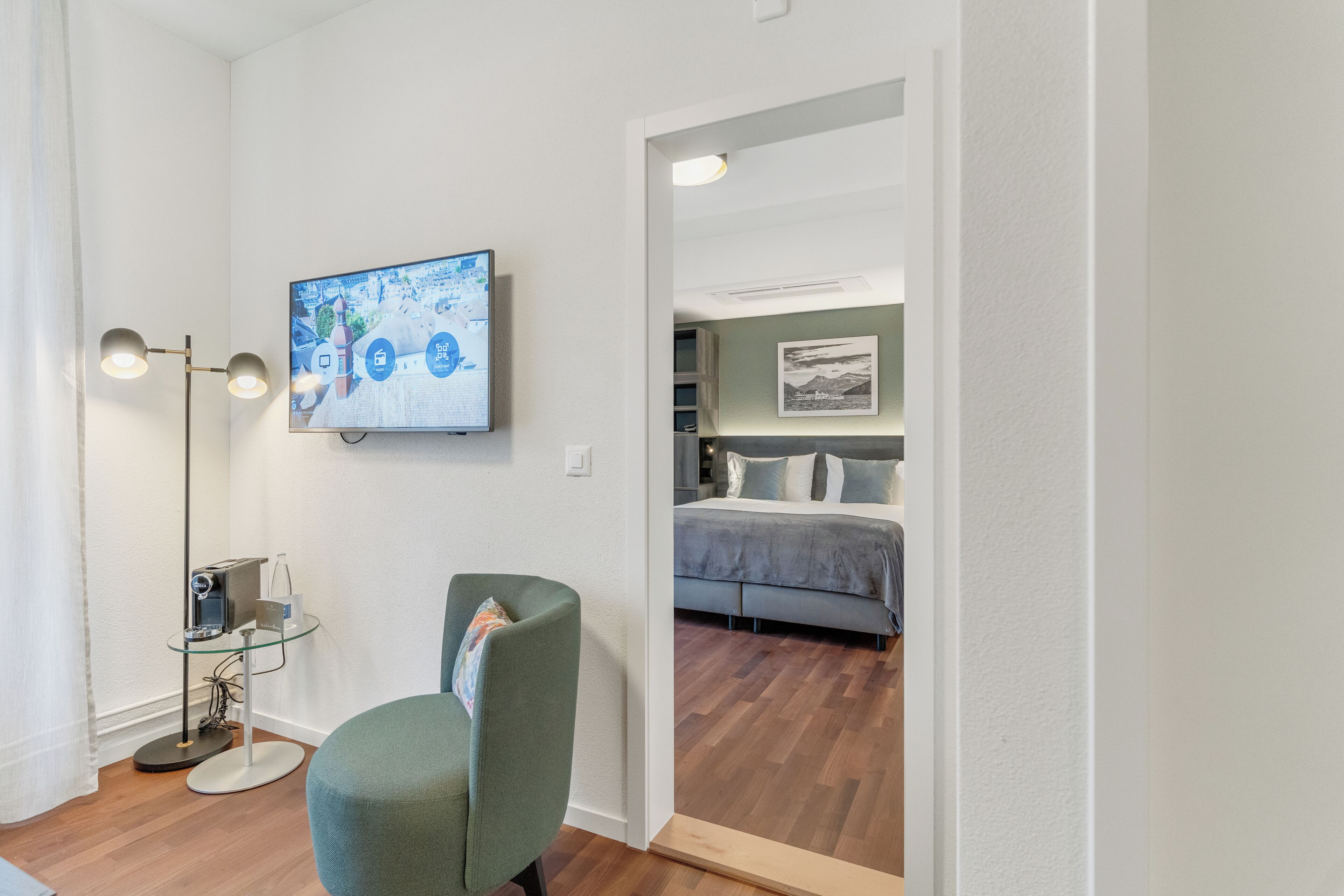 A modern room with a flat-screen TV mounted on the wall, a green armchair, a floor lamp, and a bed with a gray headboard in the adjacent room.