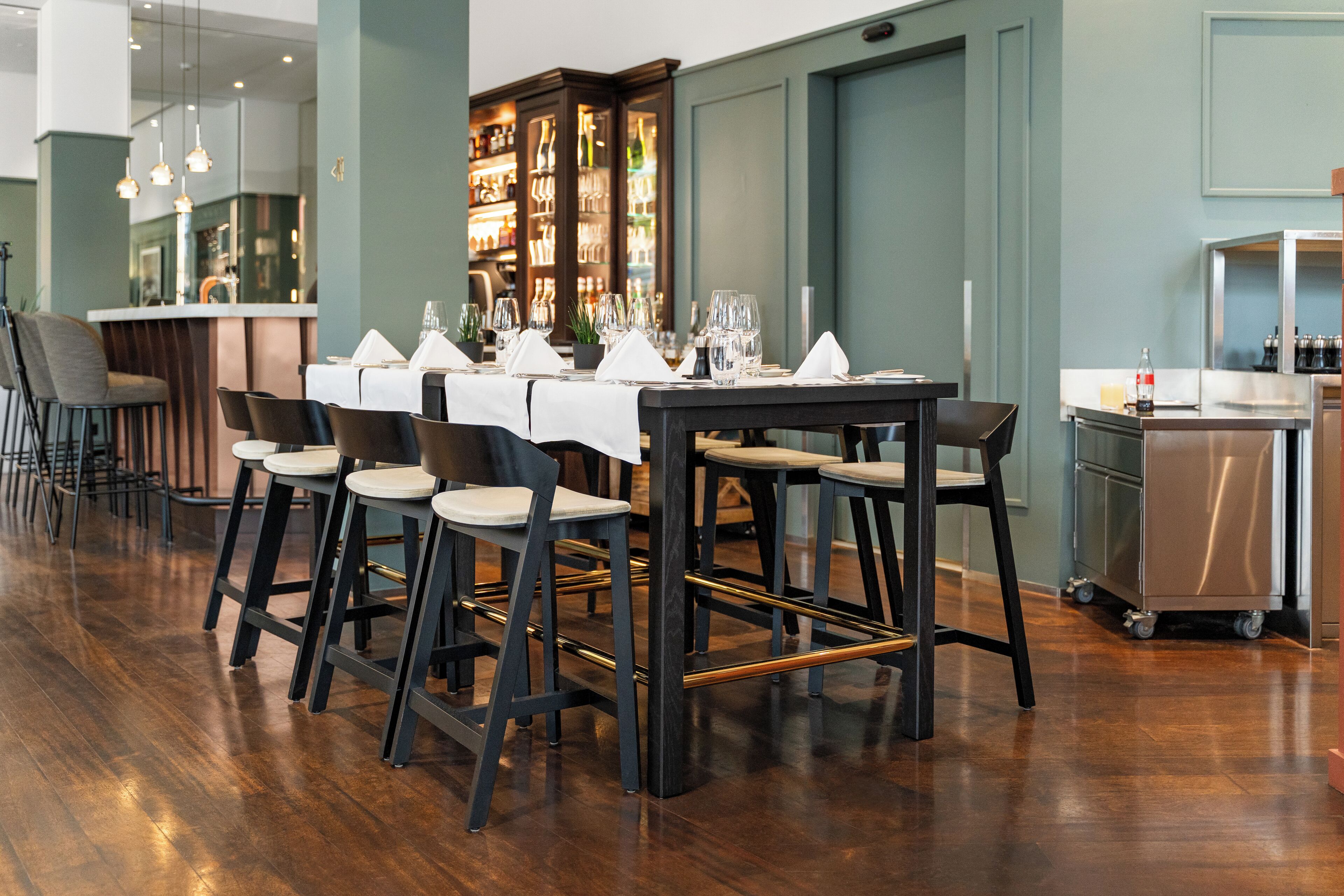 A restaurant with a long table set for dining, bar stools, and a bar area with shelves of bottles.