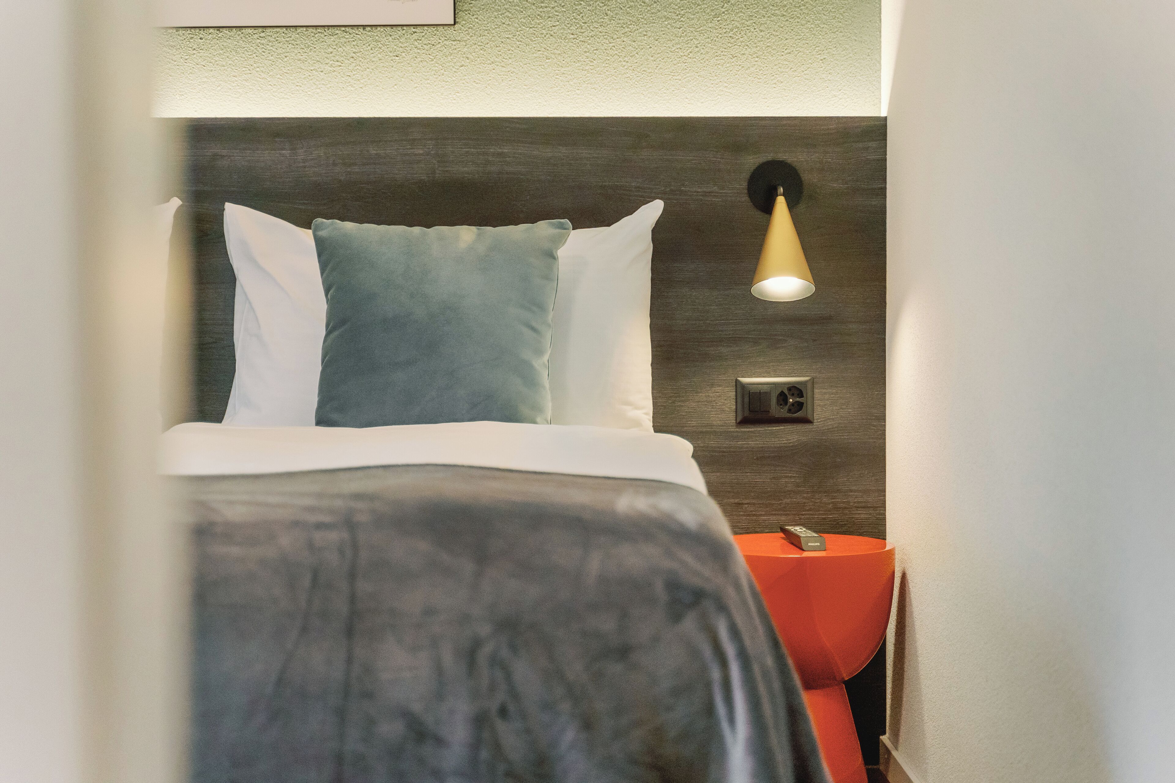 Un letto rifatto con un piumone grigio e bianco, un cuscino blu e un comodino rosso con una lampada.