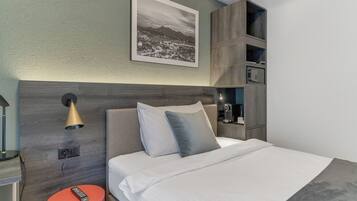 Una habitación de hotel moderna con cama, mesita de noche, lámpara, un cuadro con una vista urbana enmarcada y estanterías empotradas.