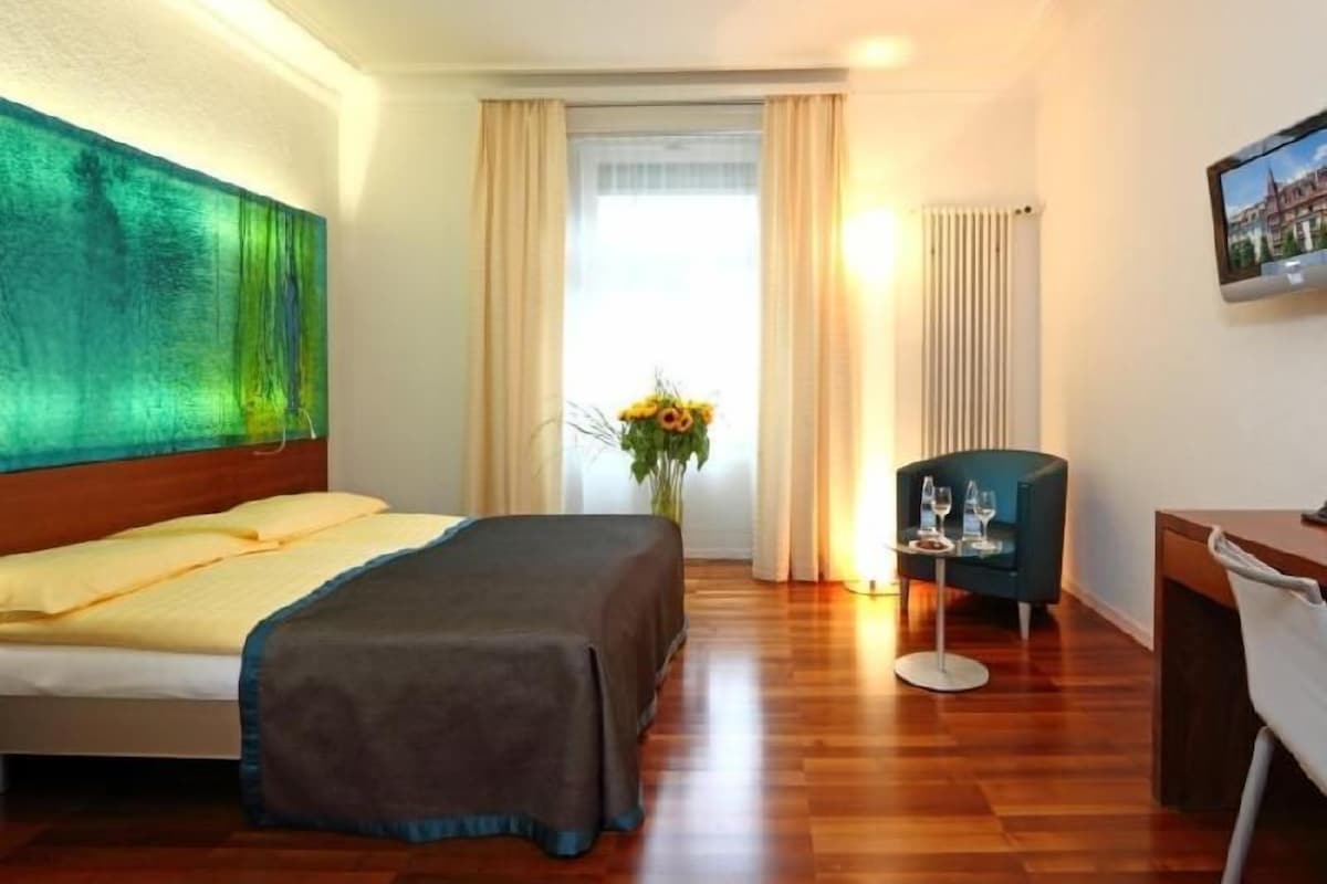 Ein Hotelzimmer mit Bett, Schreibtisch, Stuhl, Blumen in einer Vase und einem Bild an der Wand.