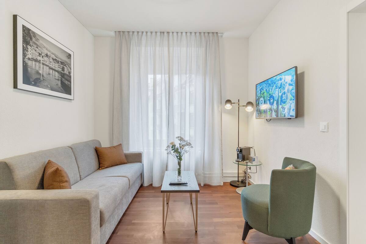 Un salon moderne comprenant un canapé, une petite table avec un vase, un fauteuil vert, un téléviseur à écran plat et une grande fenêtre ornée de rideaux légers.