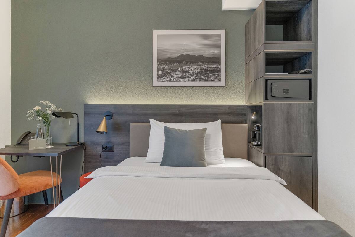Une chambre d’hôtel bien aménagée, avec un lit, un bureau doté d’une lampe, une chaise, un tableau représentant un paysage urbain encadré au mur et des rangements intégrés.