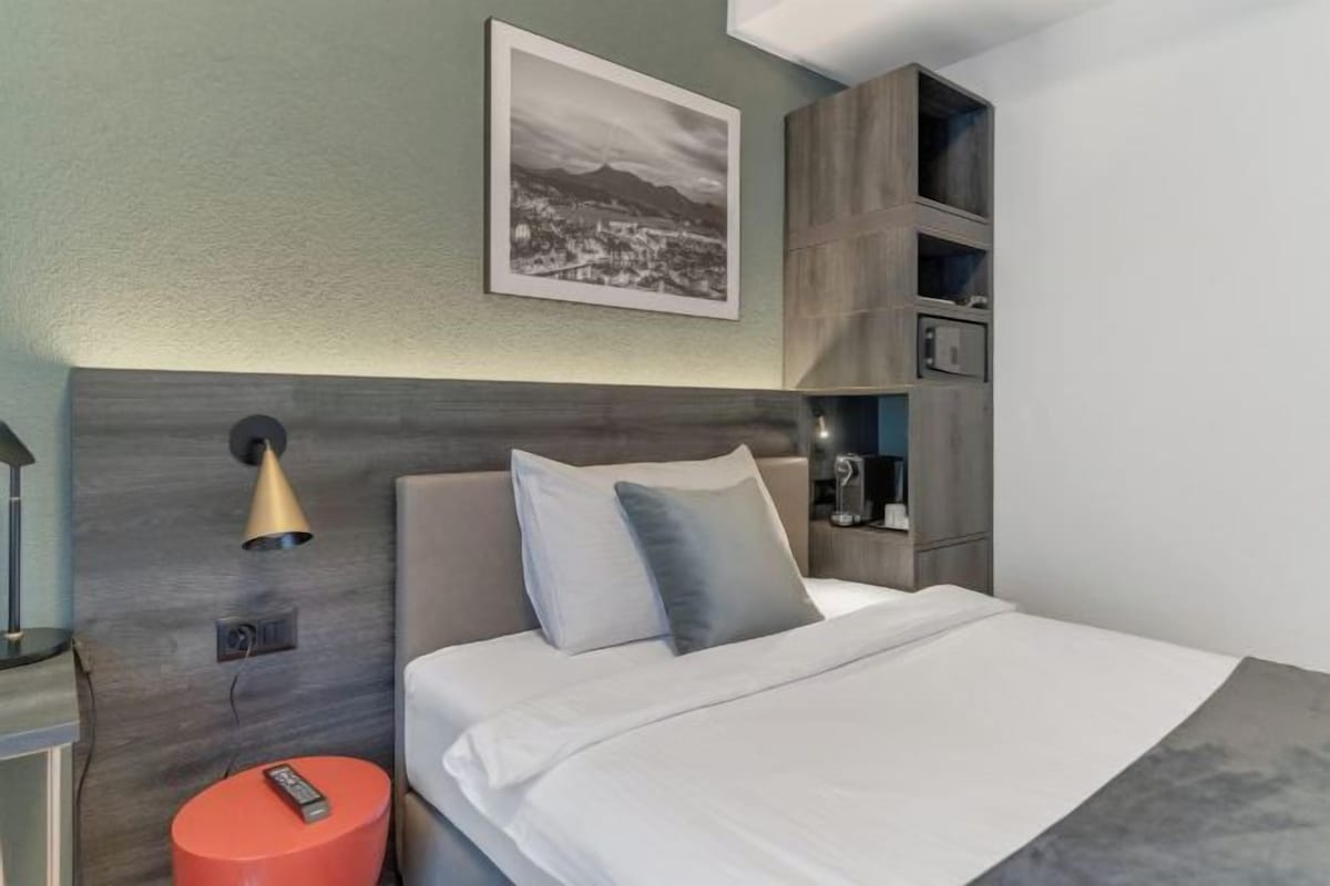 Une chambre d’hôtel moderne, équipée d’un lit, d’une table de chevet, d’un tabouret rouge, d’une lampe et d’une étagère fixée au mur.