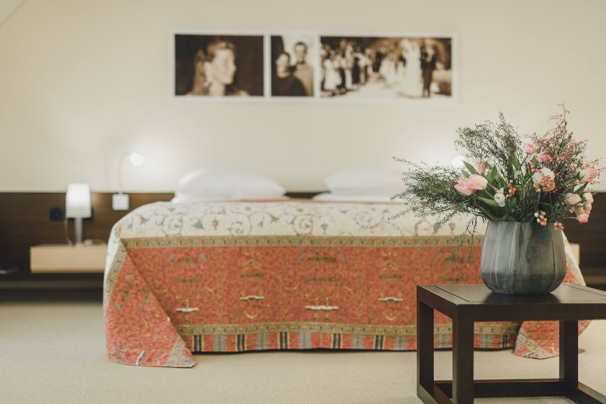 Un dormitorio con cama, una mesita de noche, un florero con flores y una pared con cuadros.