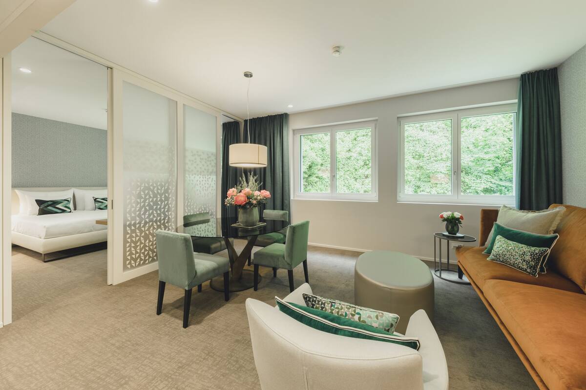 Una sala de estar moderna con un sofá, una mesa de comedor con sillas y una mesa redonda de centro. Se ven ventanas con cortinas, una lámpara y un jarrón decorativo con flores.