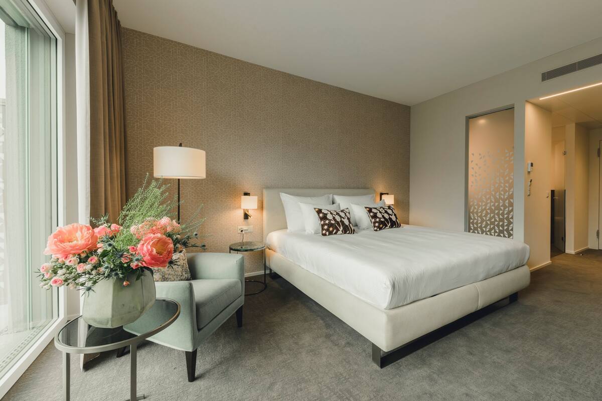Habitación de hotel con una cama grande, una zona de estar con una silla, una mesita y un jarrón de flores.
