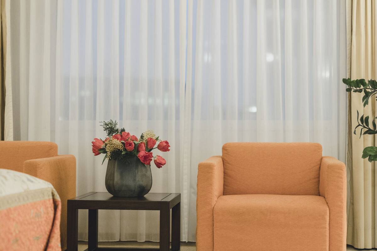 Una habitación con cama, una silla, una mesita con un florero y un ventanal con cortinas ligeras.