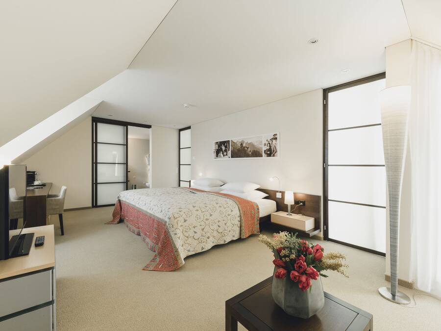 A modern hotel room with a bed, desk, and computer, featuring a large window and a vase of flowers.